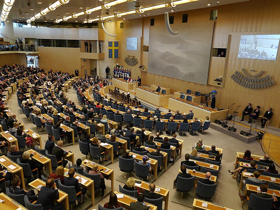 A large assembly meeting in a modern legislative chamber with numerous attendees seated in curved rows, facing a stage where speakers are addressing the audience. The chamber features a wooden interior and includes a large wall tapestry.